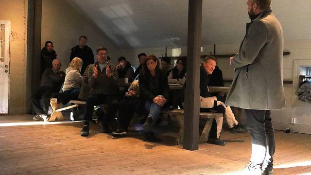 En grupp människor sitter på bänkar i ett svagt upplyst rum medan en person står och talar framför dem.