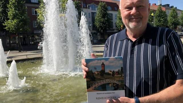 En man står framför en fontän och håller i en bok med titeln ”Kumlabygden”. I bakgrunden syns byggnader och träd.