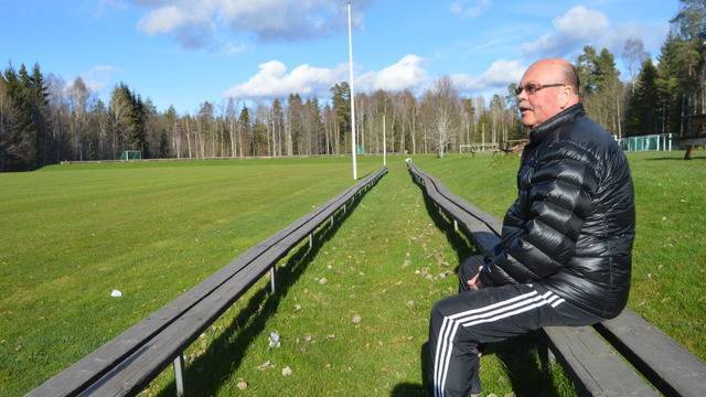 En man i svart jacka och randiga byxor sitter ensam på läktaren vid en tom gräsplan, med träd och blå himmel i bakgrunden.