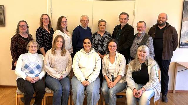 Fjorton vuxna personer poserar inomhus i två rader, några sittande och andra stående, mot en krämfärgad vägg i ett väl upplyst rum.