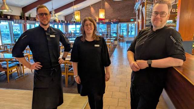 Tre restauranganställda i svarta uniformer står och ler i en rymlig, väl upplyst matsal med bord och stolar i trä.
