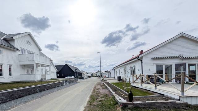 En förortsgata kantad av moderna vita hus under en molnig himmel, med en asfalterad väg och några gräsmattor i blickfånget.