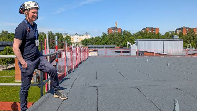En man med hjälm och skyddsutrustning står på ett platt hustak under klar himmel, med byggnader, träd och ett staket i bakgrunden.