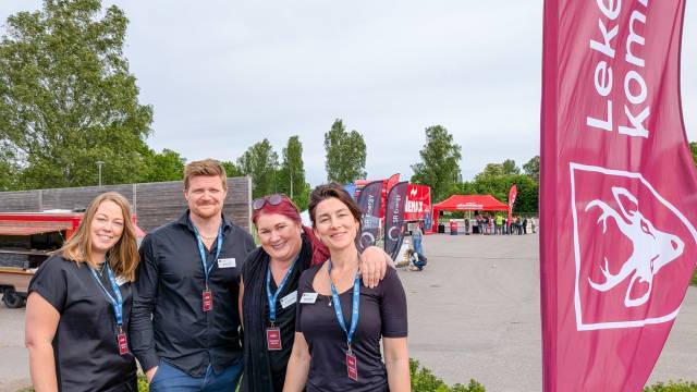 Fyra personer med namnbrickor och nyckelband står utomhus nära en stor flagga med en tjurlogotyp och text. Röda tält och träd syns i bakgrunden.