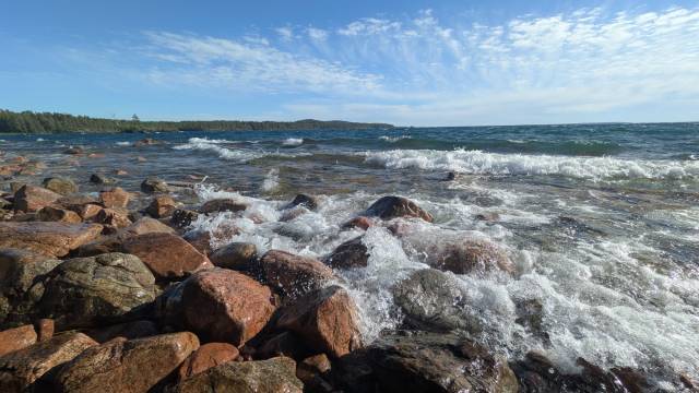 Vågorna slår mot rödaktiga klippor längs en stenig kust under en blå himmel med spridda moln; i fjärran syns skogsmark.