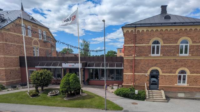 En tegelbyggnad med svart plåttak, en flaggstång med vit flagga och en huvudentré omgiven av buskar och träd under en delvis molnig himmel.