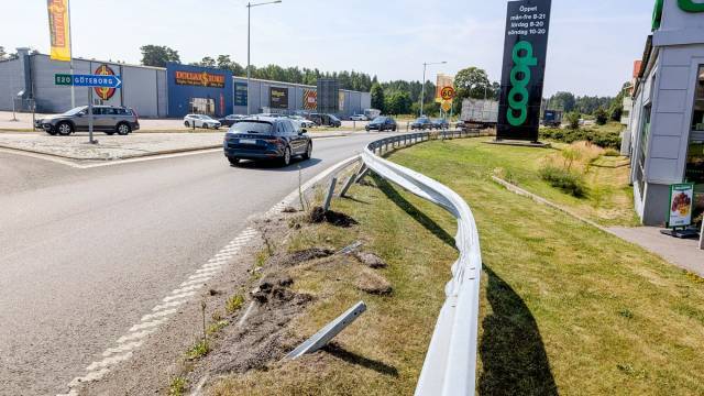 Ett skadat vägräcke med böjda delar löper längs en trafikerad gata nära butiker, en parkeringsplats och en skylt som visar Coop-supermarknadens öppettider.