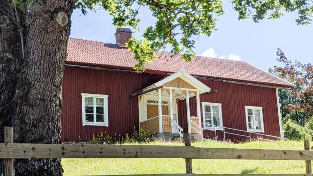 Ett rött trähus med en veranda med vita lister ligger bakom ett trästaket och ett stort träd en solig dag.