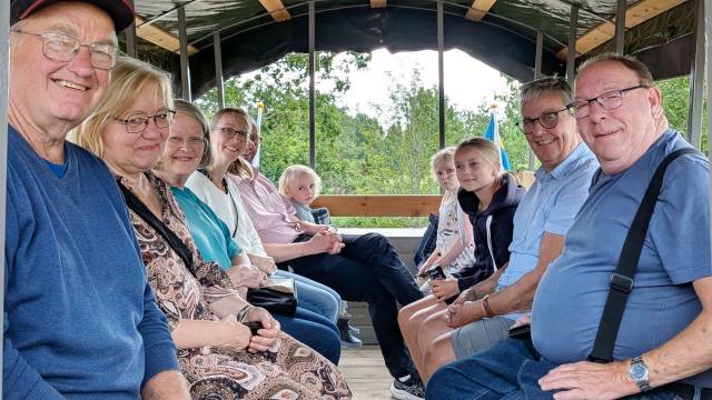 En grupp vuxna och barn sitter mittemot varandra på träbänkar inuti en täckt vagn och ler mot kameran under en utflykt på dagen.