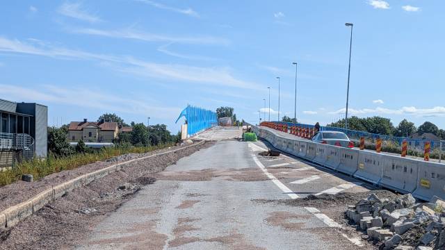 En delvis färdigställd väg med byggstaket, byggavfall och ett fordon i fjärran. Blå stängsel kantar den ofärdiga sträckan under en klar himmel.
