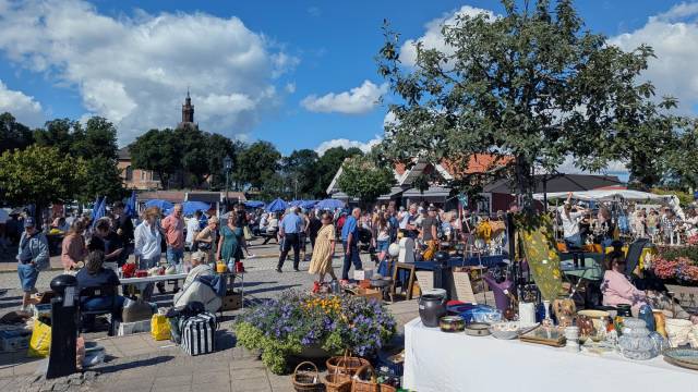Folk strosar bland stånden och borden med olika varor på en utomhusmarknad en solig dag, med träd, blommor och en kyrka synliga i bakgrunden.