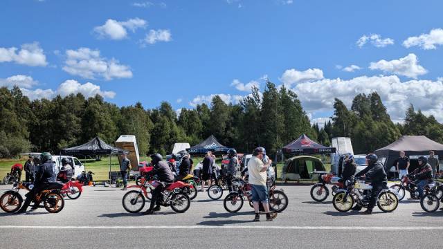 Människor på små motorcyklar står uppställda på en väg nära flera tält, med åskådare och träd i bakgrunden under en delvis molnig himmel.