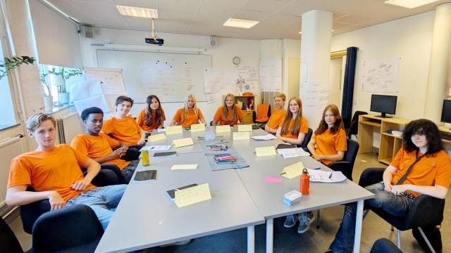 Tolv ungdomar i orangea tröjor sitter runt ett bord i ett klassrum med papper, namnskyltar och tuschpennor, vända mot kameran.