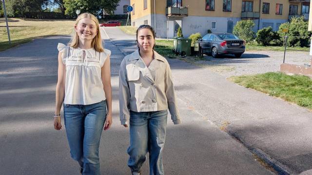 Två unga kvinnor går sida vid sida på en bostadsgata i dagsljus; båda har på sig jeans och ljusa överdelar.