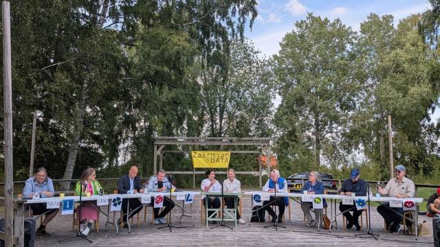Elva personer sitter vid bord med mikrofoner och partiskylter utomhus under träden och deltar i en paneldiskussion. I bakgrunden hänger en gul banderoll med texten ”Zakrisson DATA”.