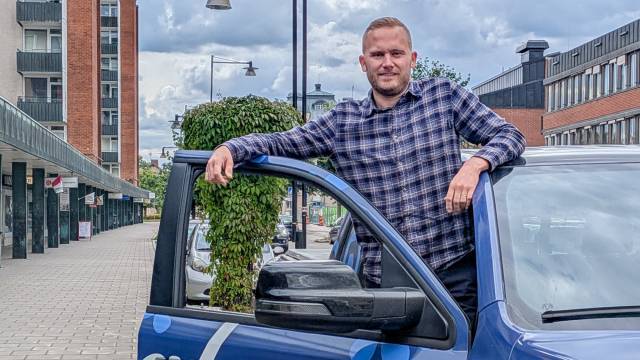 En man i en rutig skjorta står bredvid en öppen bildörr på en gata i staden, med byggnader och parkerade bilar synliga i bakgrunden.