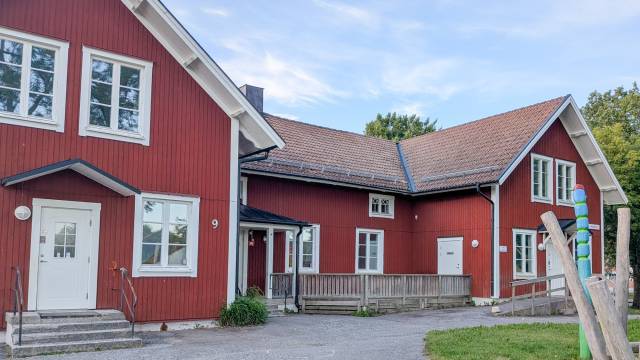 En röd träbyggnad med vita lister, flera fönster, ett sadeltak och en liten ramp som leder till ingången. I bakgrunden syns träd och en färgglad stolpe.