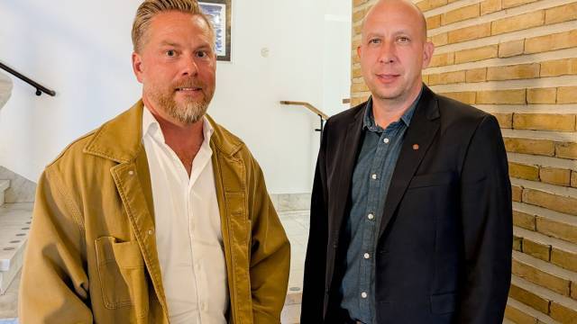 Två män står inomhus nära en trappa och en tegelvägg, båda vända mot kameran; den ene bär en beige jacka, den andre en mörk kavaj.