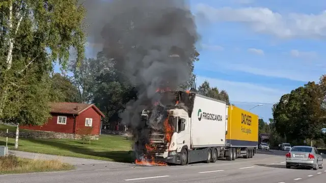 En stor vit lastbil, vars förarhytt står i lågor, avger tjock svart rök medan den står parkerad vid sidan av vägen; i närheten syns ett rött hus och förbipasserande bilar.