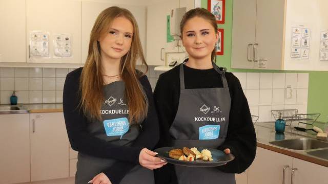 Två unga kvinnor i förkläden står i ett kök, håller i en tallrik med mat och tittar in i kameran.