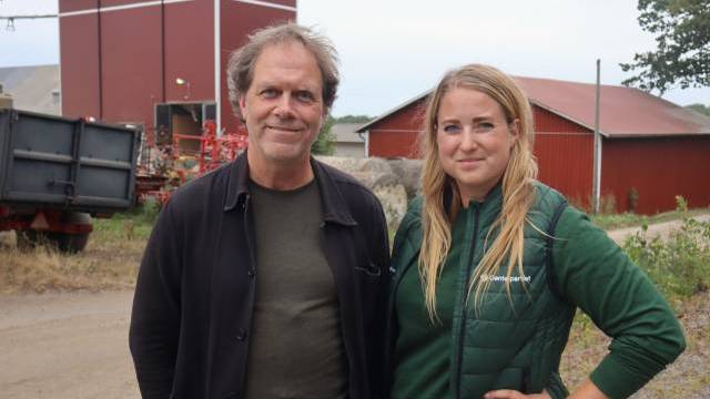 Två personer står utomhus på en grusväg framför röda lantbruksbyggnader och poserar för kameran. Det verkar vara en lantlig miljö.