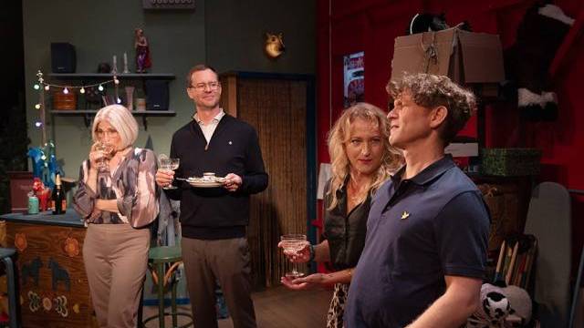 Fyra vuxna står i ett färgglatt, rörigt rum, med drinkar och snacks i händerna, och verkar konversera och umgås i en avslappnad inomhusmiljö.