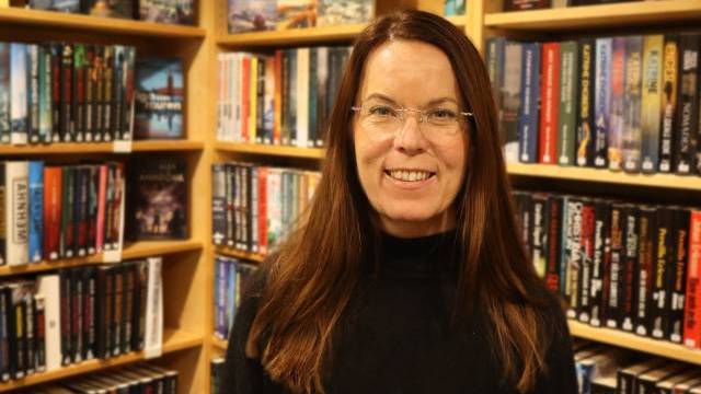 En kvinna med långt brunt hår och glasögon står leende framför bokhyllor fyllda med olika böcker på ett bibliotek eller i en bokhandel.