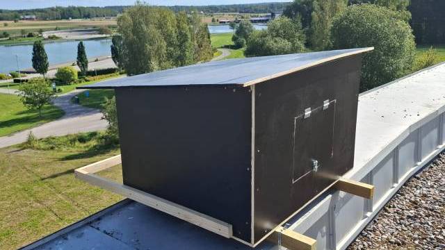 En stor svart trälåda med en liten dörr är monterad på ett hustak med utsikt över ett grönt landskap och en vattensamling i fjärran.