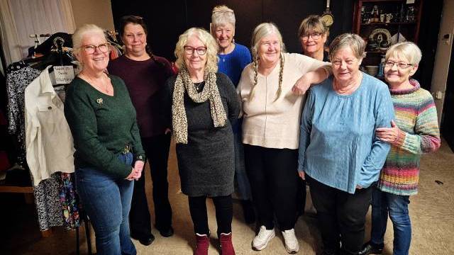 Åtta äldre kvinnor står tillsammans inomhus och ler mot kameran. Klädhängare och hyllor syns i bakgrunden. Miljön verkar avslappnad och vänlig.
