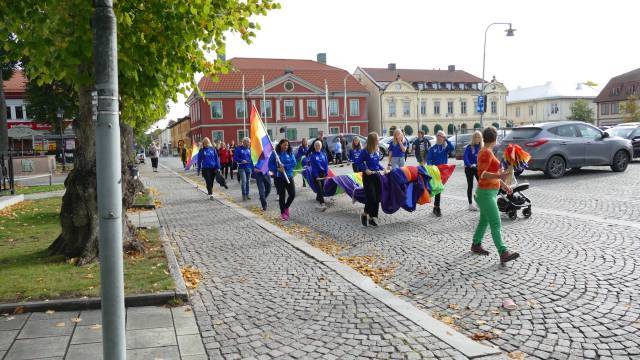 En grupp människor går nedför en kullerstensgata i en parad, bärande regnbågsflaggor, några bär blå jackor medan en person skjuter en barnvagn.