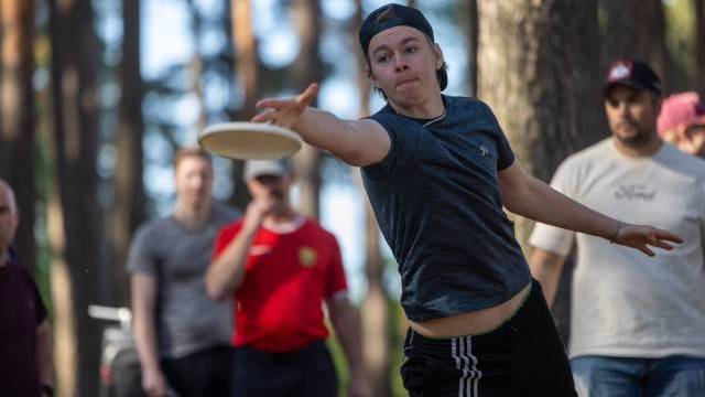 En ung person i svart keps och t-shirt kastar en disc i en skogig utomhusmiljö, medan flera personer tittar på i bakgrunden.