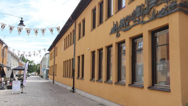 En kullerstensgata med gula hus, uteserveringar, dekorativa metallkonstverk och hängande flaggor med texten ”TÖKAGATAN”.