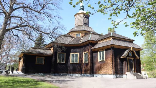 En träkyrka med ett sexkantigt torn och flera spetsiga tak, omgiven av träd och en gräsyta en solig dag.