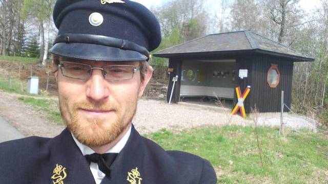 En man i mörk uniform och keps tar en selfie utomhus nära en liten svart byggnad med ett glasfönster och en färgglad stolpe. Träd och gräs syns i bakgrunden.