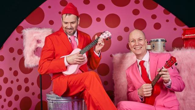 Två män i ljusröda och rosa kostymer spelar ukuleles på en rosa prickig bakgrund, sittande på rosa pälsstolar med musikinstrument i närheten.