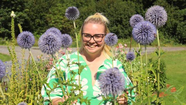 En person med glasögon och en grön blommig skjorta står och ler bland höga, lila alliumblommor i en solig trädgård.