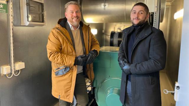 Två män i rockar och handskar står inomhus bredvid industriutrustning och ler mot kameran.