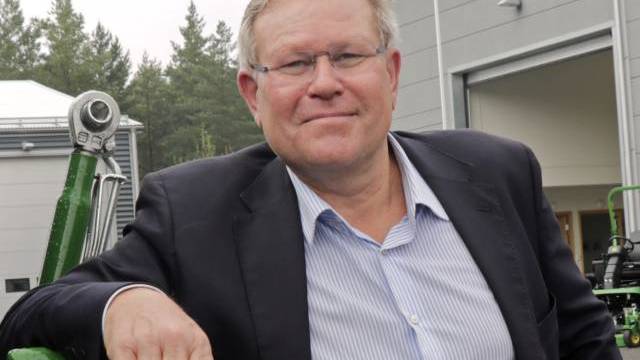 En man i kostym och glasögon står utomhus och lutar sig mot en grön maskin framför en modern industribyggnad, med träd i bakgrunden.