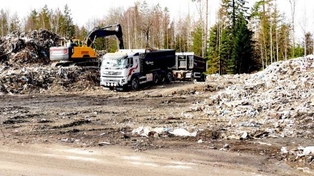 En stor lastbil och en grävmaskin arbetar bland avfallshögar på en utomhusdeponi, med träd synliga i bakgrunden.