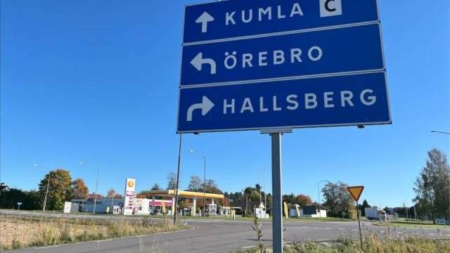 Blå vägskylt med vägbeskrivning till Kumla, Örebro och Hallsberg i en korsning nära en Shellmack under klar himmel.