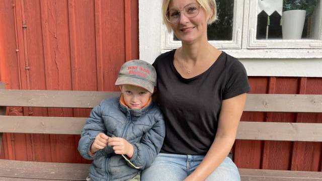 En kvinna och en ung pojke sitter på en träbänk framför en röd träbyggnad med vita fönsterkarmar.