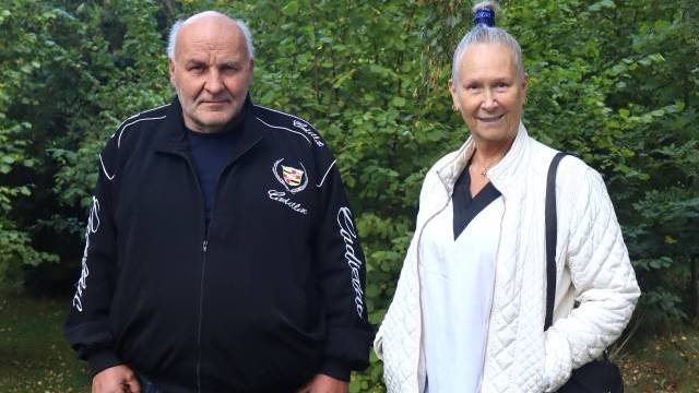 En äldre man och kvinna står utomhus framför grönt lövverk. Mannen bär en svart jacka, och kvinnan bär en vit vadderad jacka och har håret uppsatt i en knut.