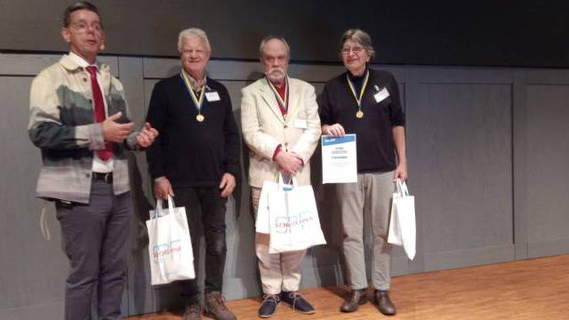 Fyra personer står på en scen; tre bär medaljer och håller i presentpåsar, medan den fjärde, i mönstrad jacka, står bredvid och talar eller presenterar.