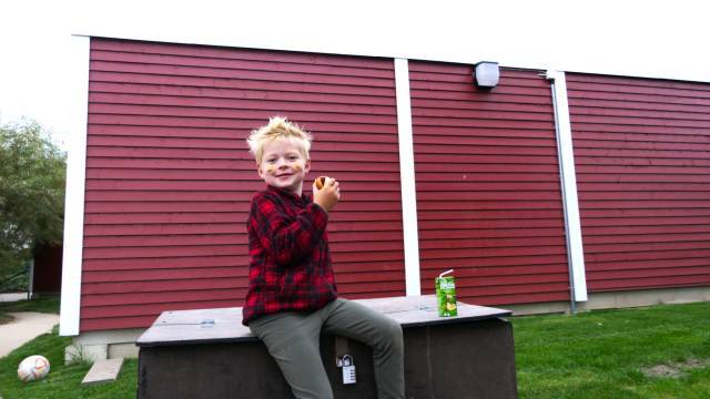 Ett barn i rödrutig skjorta sitter på en svart låda utomhus och håller i ett äpple. En juiceburk och en fotboll finns i närheten. Röd träbyggnad med vit kant i bakgrunden.