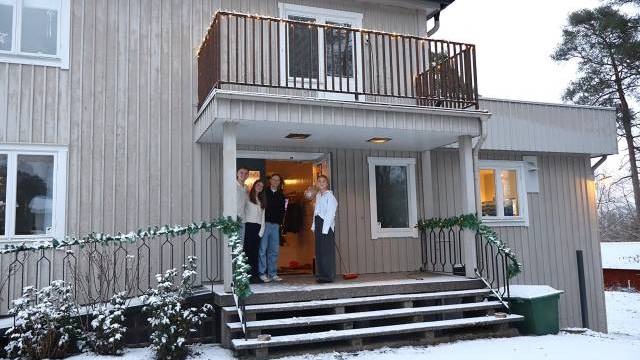 Tre personer står på verandan till ett tvåvånings trähus med snö som täcker marken och trappstegen.