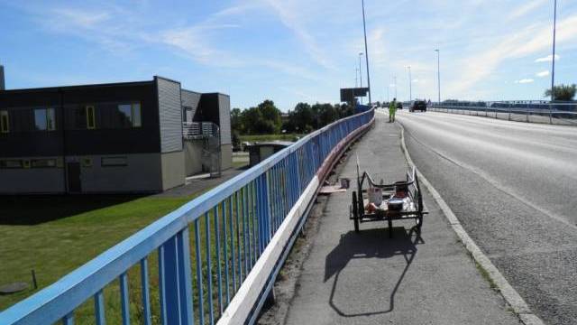 En arbetsvagn med verktyg står parkerad på trottoaren bredvid en bro med blå räcke; en arbetare i gul väst går längre fram.