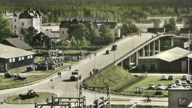 En svängd bro med bilar går över en väg och järnväg i en liten stad med träd, byggnader och parkerade fordon i bakgrunden.