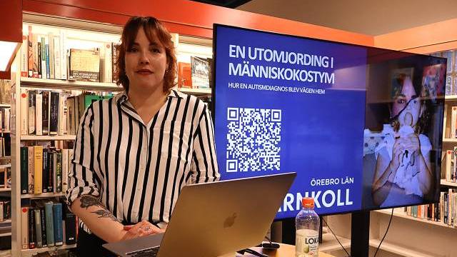 En kvinna står bredvid en bärbar dator och en presentationsskärm i ett bibliotek. På skärmen visas svensk text, en QR-kod och en bild av en person i kostym.