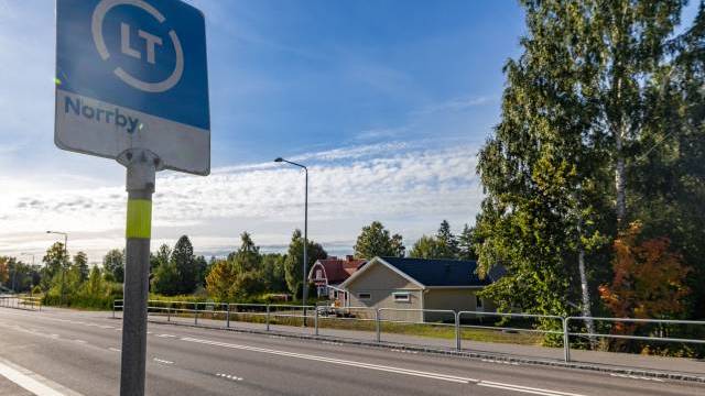 En busshållplatsskylt vid vägen med texten ”Norrby”, med träd, hus och en klarblå himmel i bakgrunden.