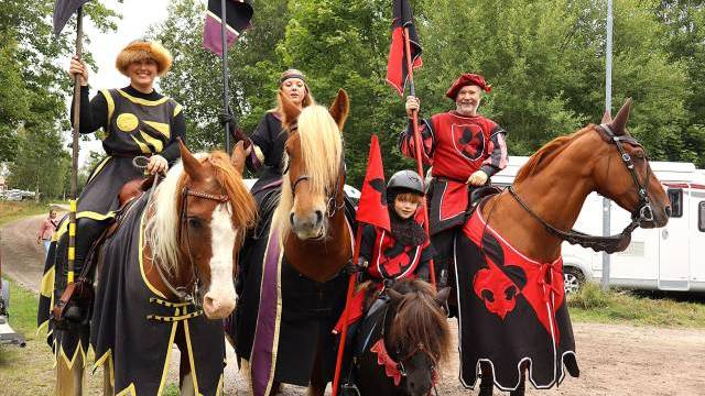 Fem personer i medeltida dräkter rider på tre hästar och en ponny med flaggor i händerna, med träd och en släpvagn i bakgrunden.